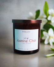 Load image into Gallery viewer, Infusion Candle Co. Jasmine Chai soy candle in amber glass jar with copper lid, set against a backdrop of white jasmine flowers and green leaves. The candle label reads 'Jasmine Chai, Floral Tea Blend' and highlights the handcrafted nature of the candle, made in Illinois. This soy wax candle blends the soothing scent of jasmine with the rich, spicy aroma of chai, perfect for creating a calming and aromatic atmosphere.
