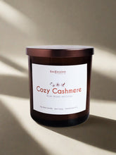 Load image into Gallery viewer, Infusion Candle Co. Cozy Cashmere soy candle in an amber glass jar with a copper lid, positioned on a beige surface with soft shadows. The candle label reads 'Cozy Cashmere, Plum, Peony, Patchouli,' highlighting the luxurious and comforting fragrance blend. This handcrafted 9 oz candle, made in Illinois, combines the rich scents of plum, peony, and patchouli, perfect for creating a warm and inviting atmosphere, ideal for enhancing home comfort and relaxation