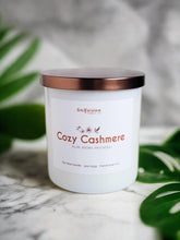 Load image into Gallery viewer, Infusion Candle Co. Cozy Cashmere soy candle in a white glass jar with a copper lid, placed on a marble surface with green leaves in the background. The candle label reads 'Cozy Cashmere, Plum, Peony, Patchouli,' highlighting the luxurious and soothing fragrance blend. This handcrafted 9 oz candle, made in Illinois, combines the rich and inviting scents of plum, peony, and patchouli, perfect for creating a warm, cozy, and relaxing atmosphere in any home