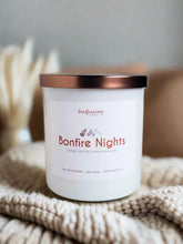 Load image into Gallery viewer, Infusion Candle Co. Bonfire Nights soy candle in a white glass jar with a copper lid, placed on a cozy knitted blanket. The candle label reads 'Bonfire Nights, Cedar, Smoke, Marshmallow,' highlighting the rustic and inviting fragrance blend. This handcrafted 9 oz candle, made in Illinois, captures the essence of a cozy bonfire night with its warm and comforting scents, perfect for creating a relaxing and nostalgic atmosphere