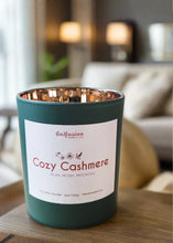 Load image into Gallery viewer, Infusion Candle Co. Cozy Cashmere soy candle in a green glass jar with a copper interior, placed on a wooden surface in a cozy living room setting. The candle label reads 'Cozy Cashmere, Plum, Peony, Patchouli,' highlighting the elegant and luxurious fragrance blend. This handcrafted 9 oz candle, made in Illinois, combines the rich scents of plum, peony, and patchouli, perfect for creating a warm, inviting, and sophisticated atmosphere, enhancing home decor and relaxation