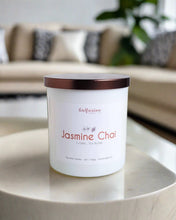 Load image into Gallery viewer, Infusion Candle Co. Jasmine Chai soy candle in a white glass jar with a copper lid, placed on a marble table in a modern living room with a beige sofa and decorative pillows in the background. The candle label features 'Jasmine Chai, Floral Tea Blend' and emphasizes the handcrafted quality of the product, made in Illinois. This soy wax candle combines the delicate fragrance of jasmine with the warm, scent of chai, ideal for enhancing relaxation and ambiance