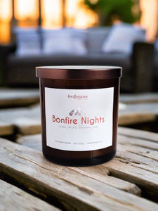 Infusion Candle Co. Bonfire Nights soy candle in an amber glass jar with a copper lid, placed on a rustic wooden surface with a blurred outdoor seating area and sunset in the background. The candle label reads 'Bonfire Nights, Cedar, Smoke, Marshmallow,' capturing the essence of a cozy evening by the fire. This handcrafted 9 oz candle, made in Illinois, combines the warm and inviting scents of cedar, smoke, and marshmallow, perfect for outdoor gatherings and cozy nights. 