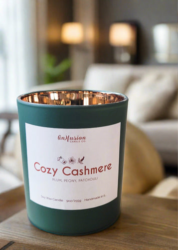 Infusion Candle Co. Cozy Cashmere soy candle in a green glass jar with a copper interior, placed on a wooden surface in a cozy living room setting. The candle label reads 'Cozy Cashmere, Plum, Peony, Patchouli,' highlighting the elegant and luxurious fragrance blend. This handcrafted 9 oz candle, made in Illinois, combines the rich scents of plum, peony, and patchouli, perfect for creating a warm, inviting, and sophisticated atmosphere, enhancing home decor and relaxation